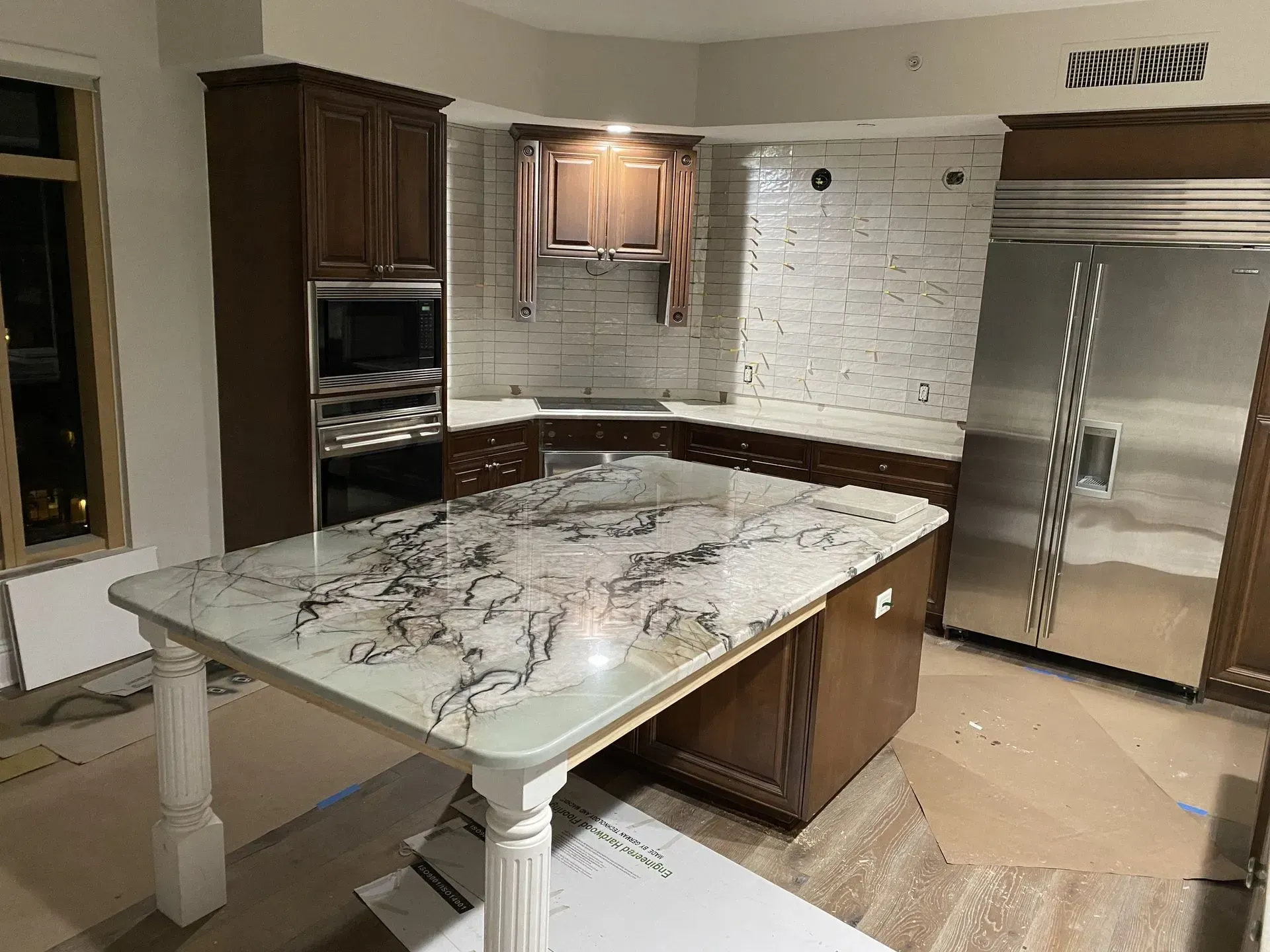 Kitchen under construction, brown cabinets, island with marble countertop, stainless steel refrigerator.