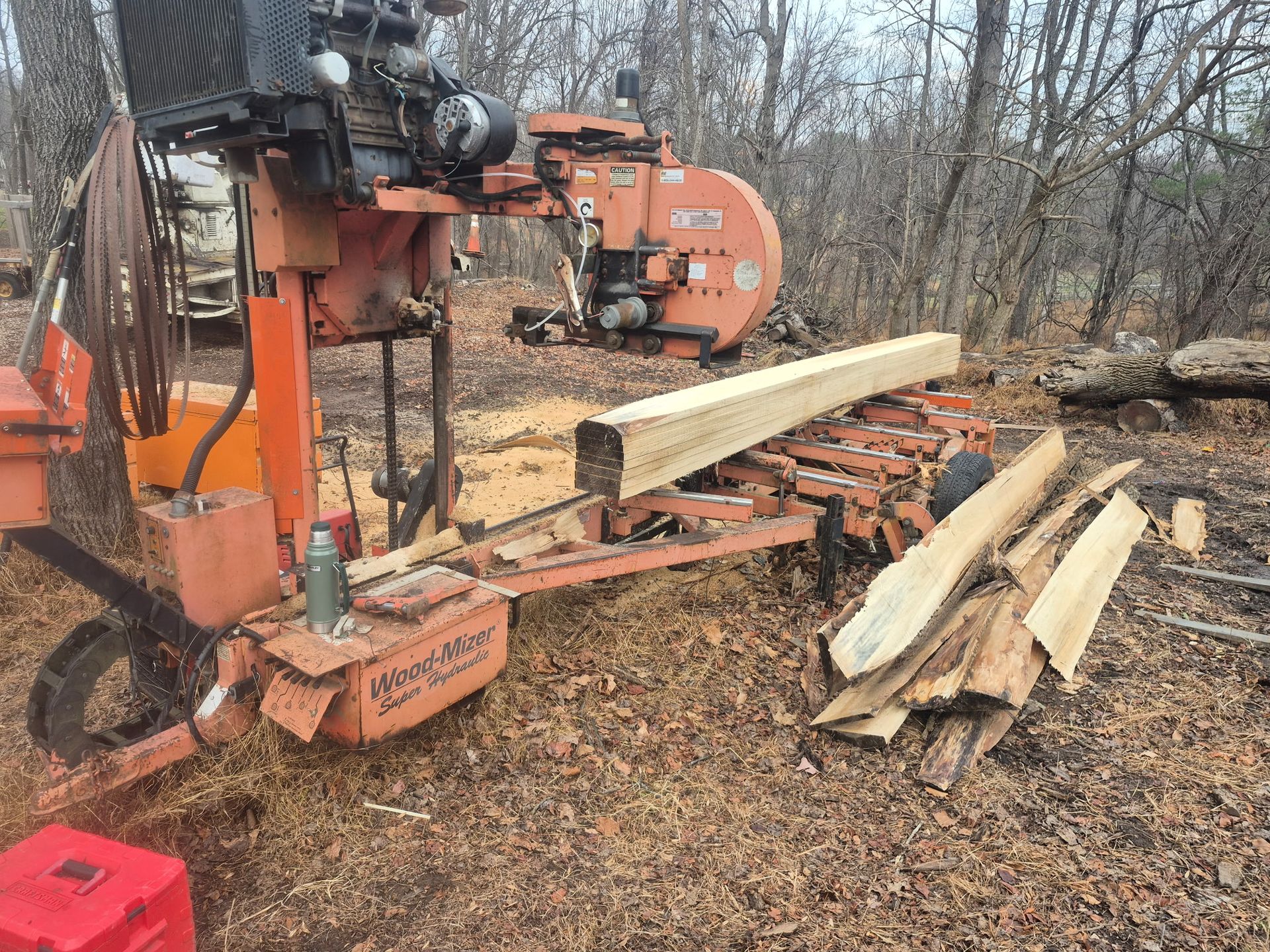 Orange sawmill cutting a log outdoors; sawdust, wood scraps, and forest setting.