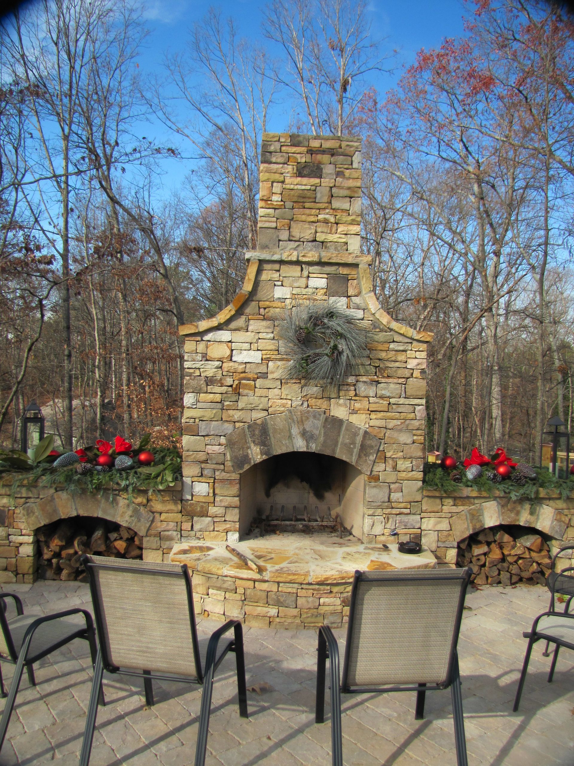 A rustic stone outdoor fireplace with two arched wood-storage alcoves, set on a patio with chairs in a wooded yard.
