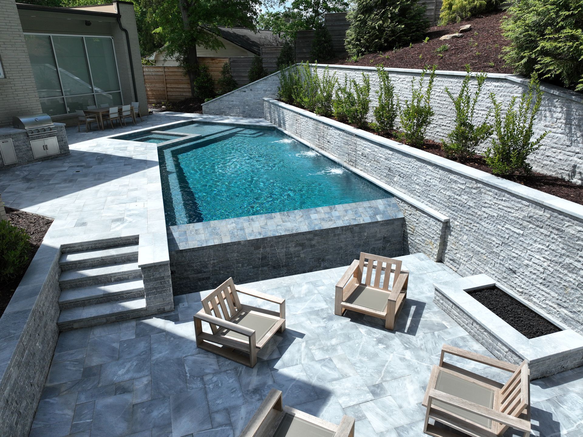A high-angle view of a backyard with a rectangular pool, gray stone patio, a fire pit, and wooden lounge chairs.