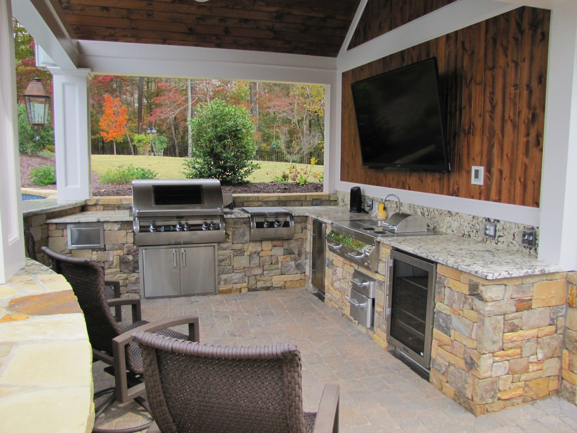 Outdoor kitchen with a stone grill island, granite countertops, bar fridge, and television under a wooden-roofed patio.