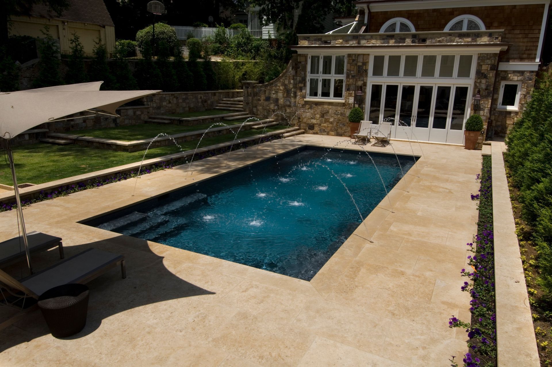A rectangular dark blue swimming pool with water fountains sits on a beige stone patio next to a stone house and garden.