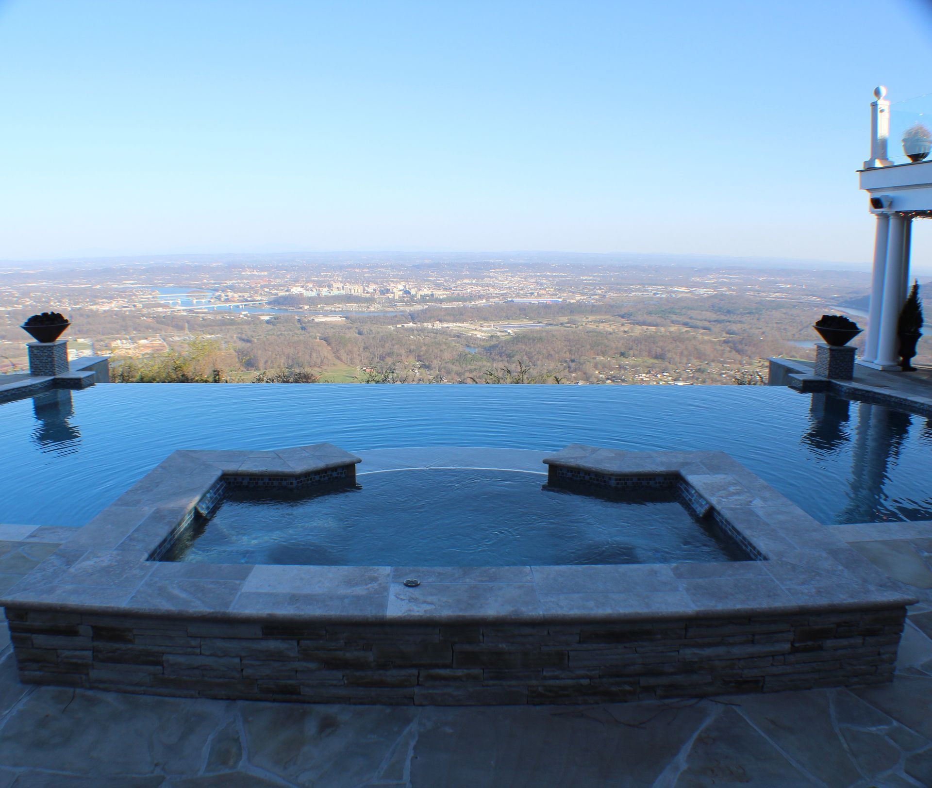An infinity pool with a built-in spa overlooks a panoramic view of a city and forested valley under a clear blue sky.