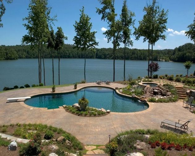 A kidney-shaped swimming pool with a diving board sits on a stone patio overlooking a large lake surrounded by trees.
