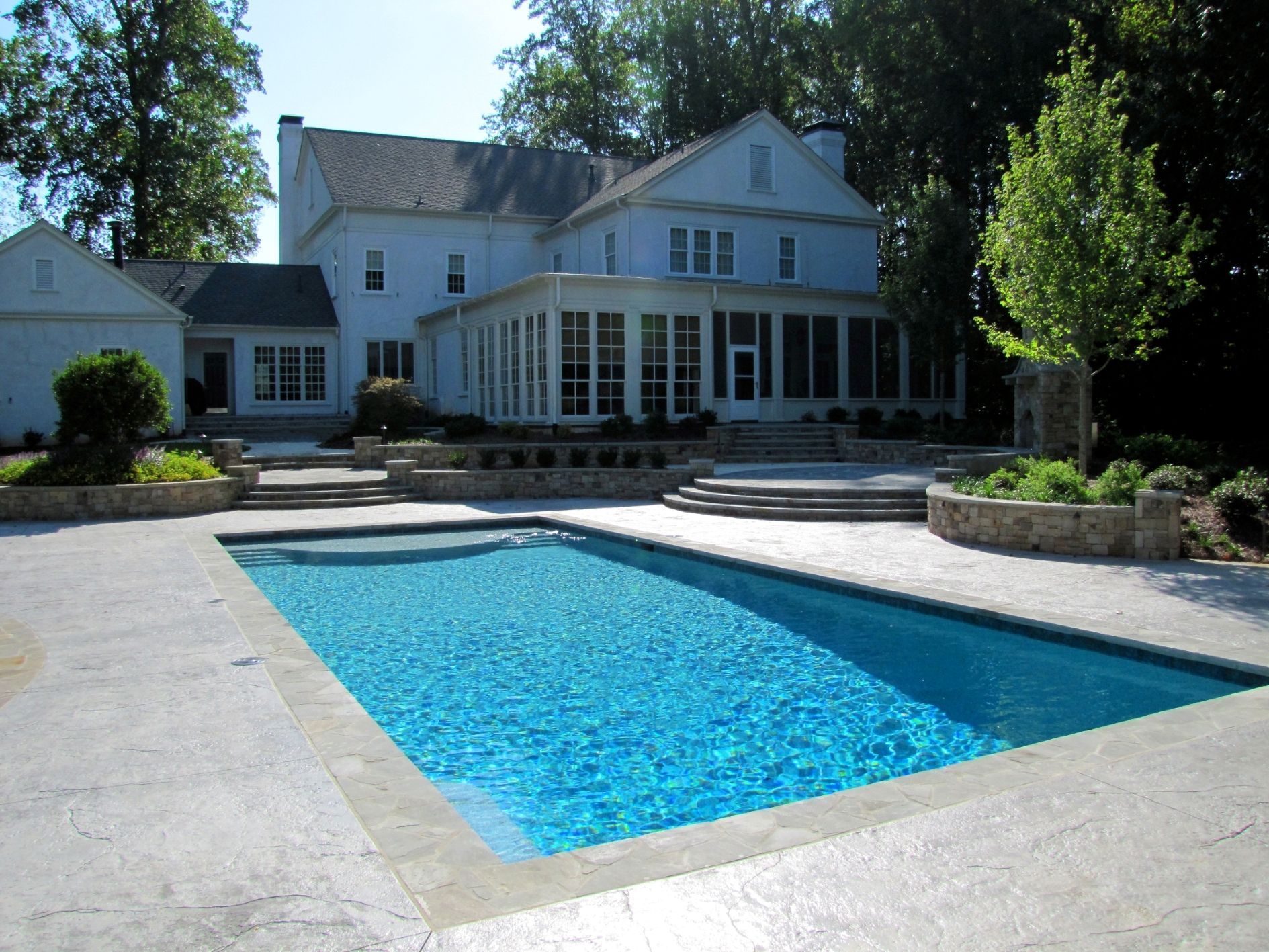 A rectangular swimming pool sits in front of a white two-story house with a stone patio and landscaped garden areas.