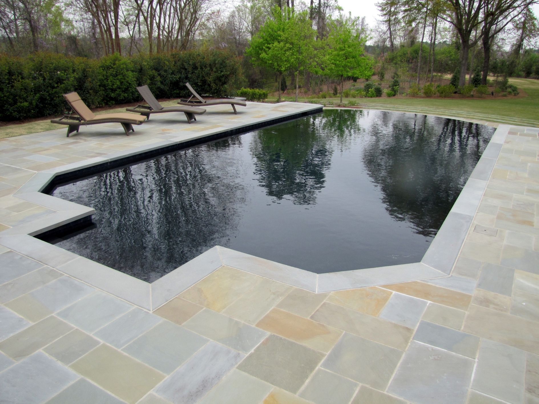 A rectangular swimming pool with dark water and a stone patio surround, featuring three lounge chairs on the deck.