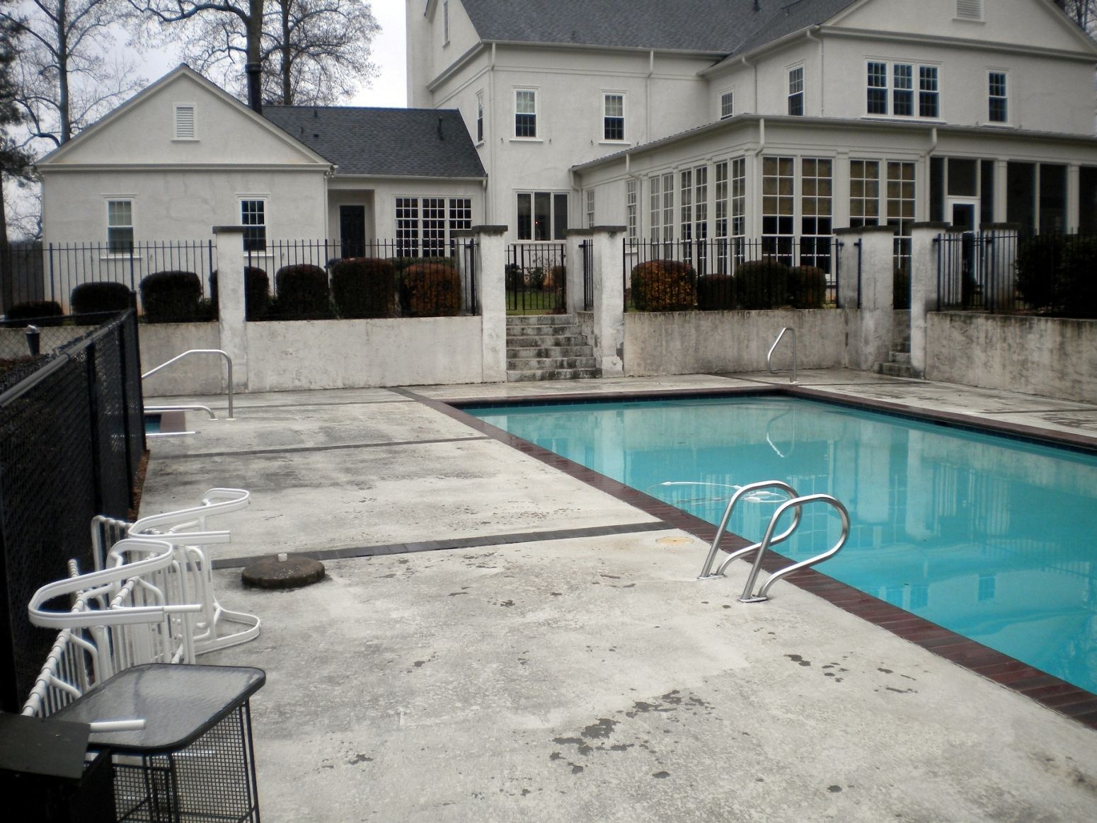An outdoor swimming pool with a white house in the background, bordered by a concrete patio and black metal fencing.