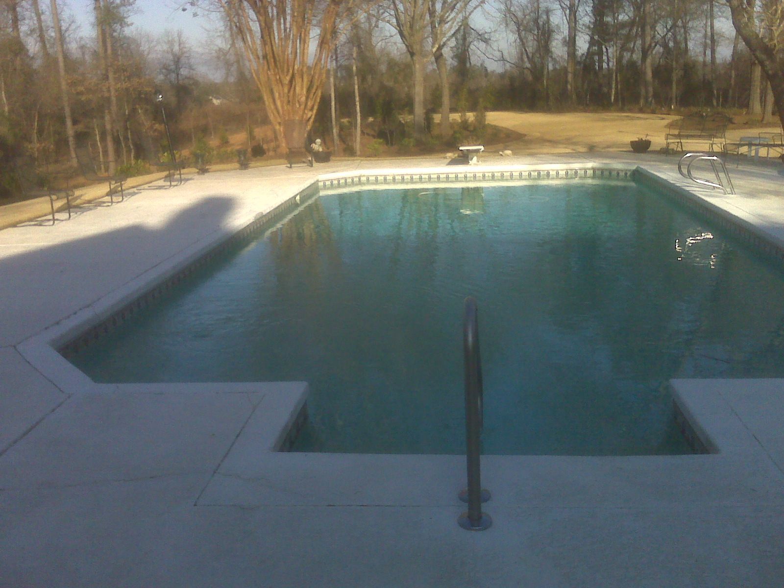 A rectangular swimming pool with a cutout corner sits on a light gray concrete deck, surrounded by trees in a grassy yard.