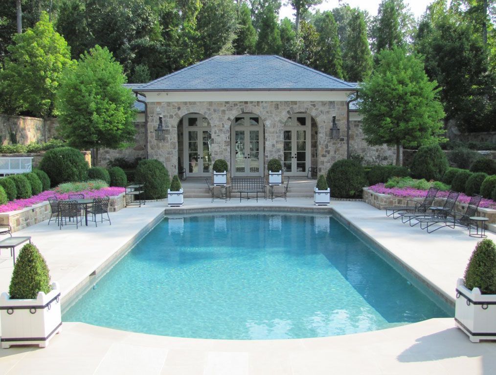 A rectangular blue swimming pool sits in front of a stone pool house, surrounded by manicured shrubs and pink flowers.