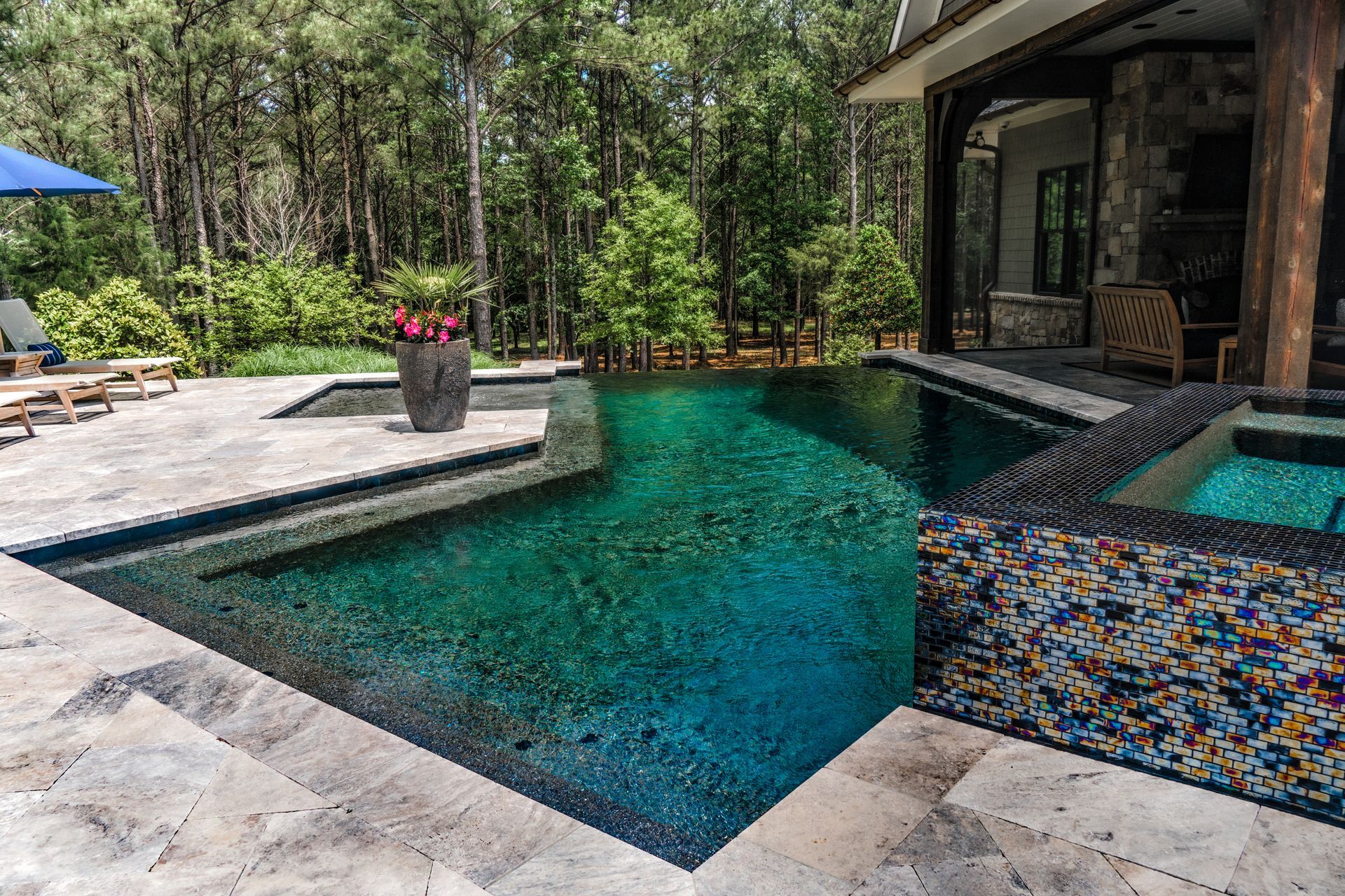 A swimming pool with dark teal water and a tiled spa sits on a stone patio surrounded by a lush forest.