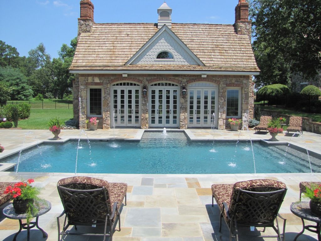 A stone pool house with a cedar roof, arched doors, and a swimming pool with fountains on a sunny patio with lounge chairs.