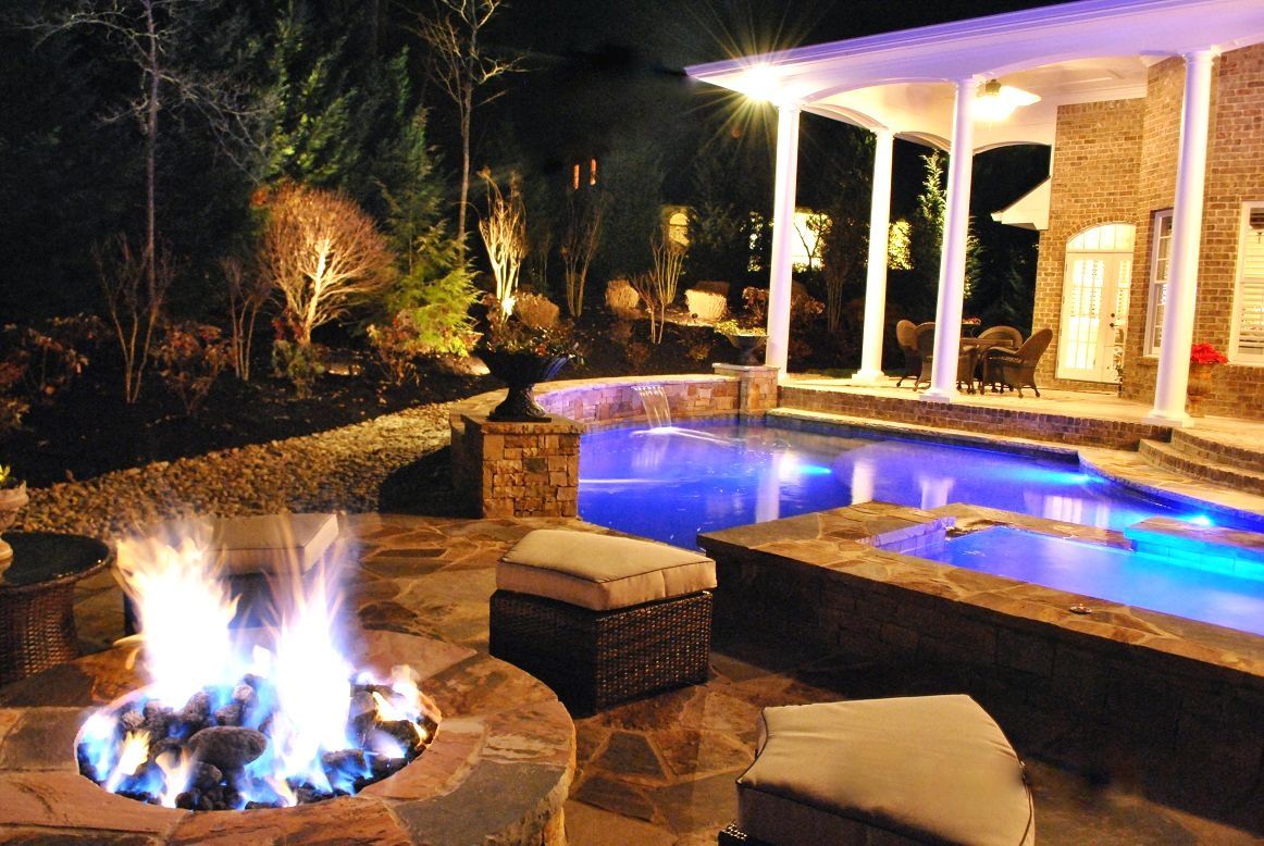 A nighttime patio view with a stone fire pit, a glowing blue swimming pool, and a white-pillared covered deck.