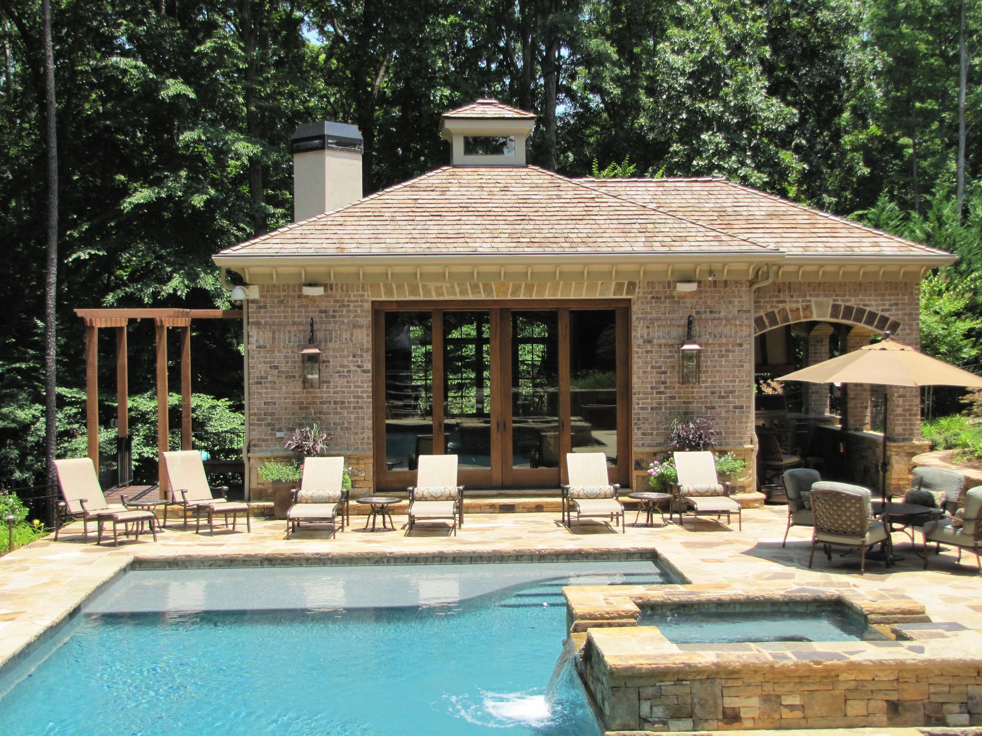 A stone pool house with a tan roof sits behind a blue swimming pool and spa on a sunny, wooded patio with lounge chairs.