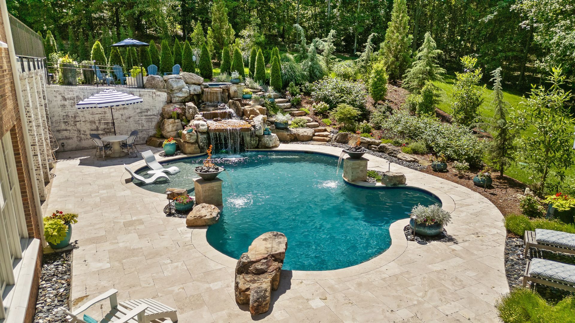 Pool area with waterfall, blue water, stone patio, lush green landscaping.