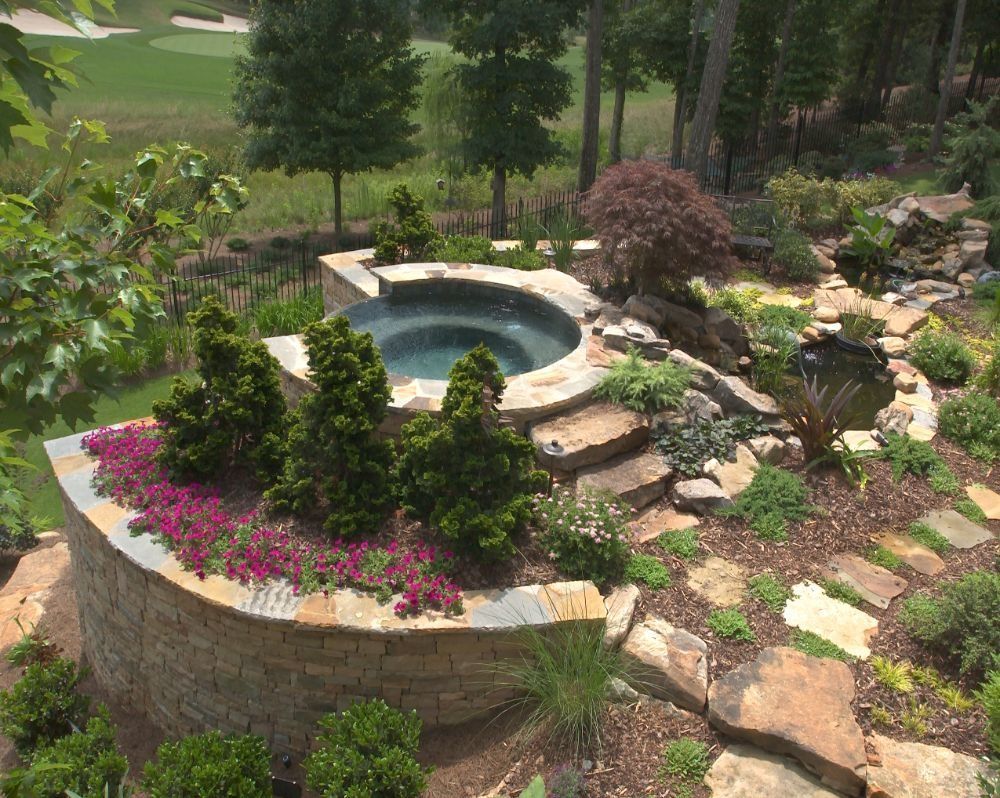 Stone-surrounded hot tub in a landscaped garden with stairs, small trees, and colorful flowers.