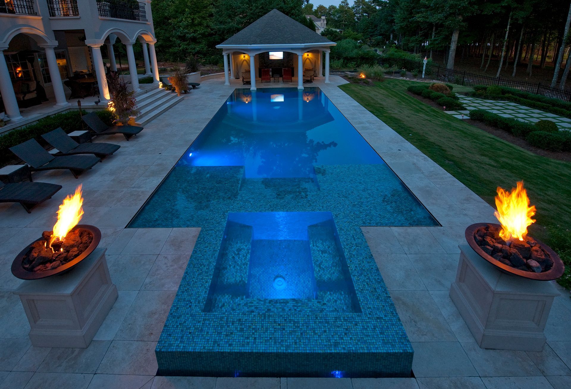 Long rectangular pool with lit water, a gazebo, and fire pits at dusk.