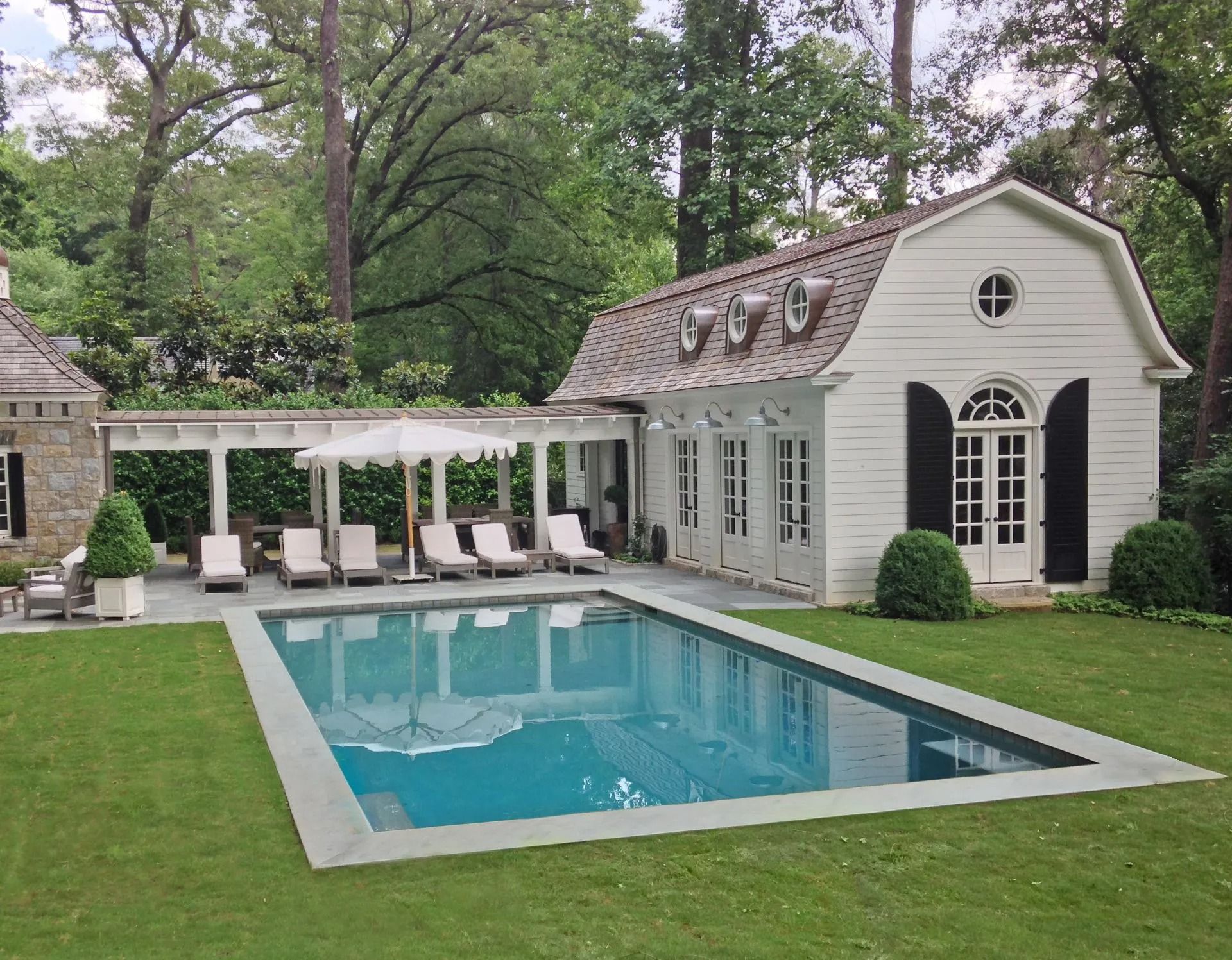 Swimming pool with lounge chairs, next to a white building with black shutters and a pergola, surrounded by a green lawn.