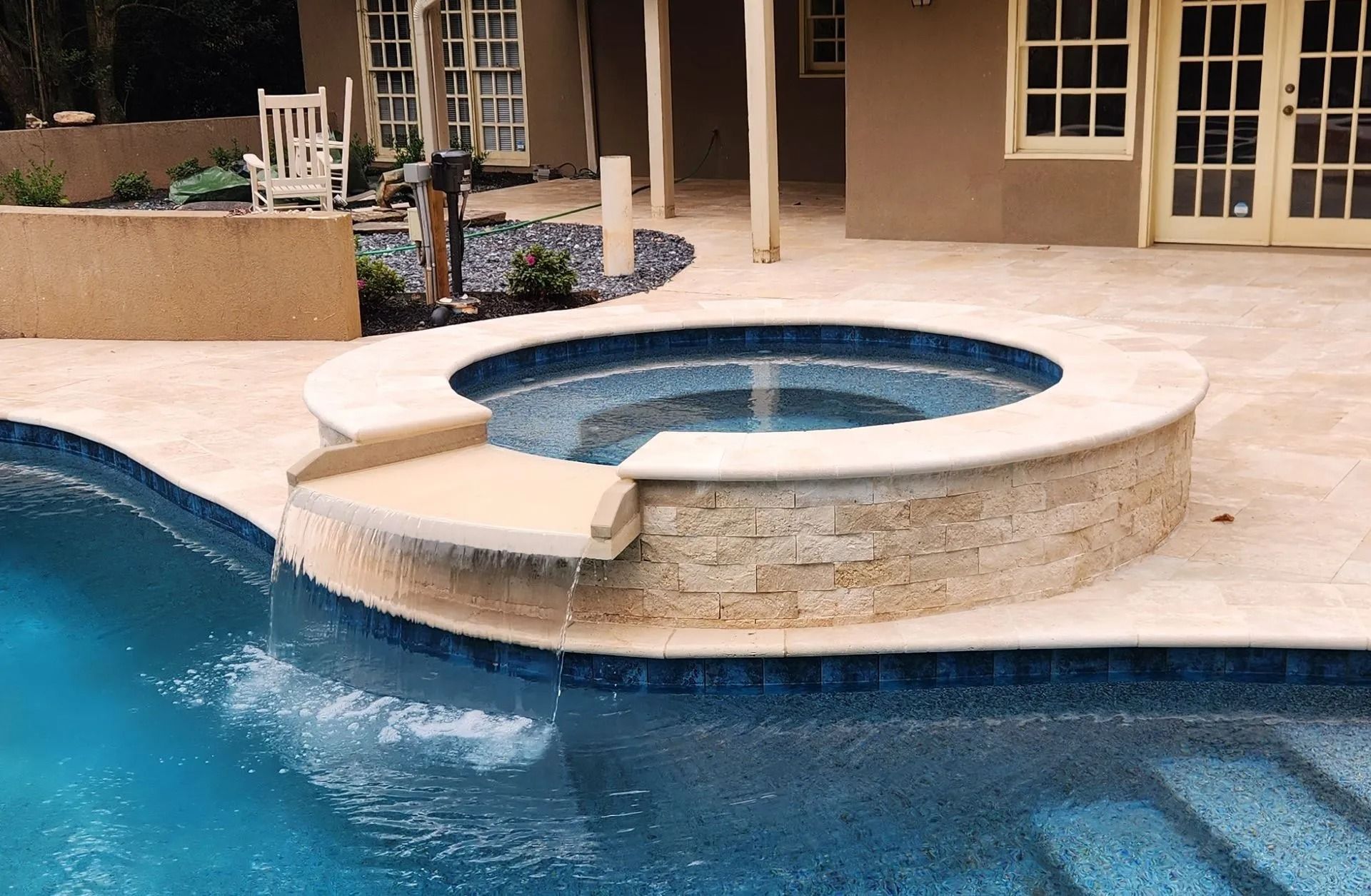 A circular hot tub with water cascading into a pool. Tan stone and blue tile. Outdoors.