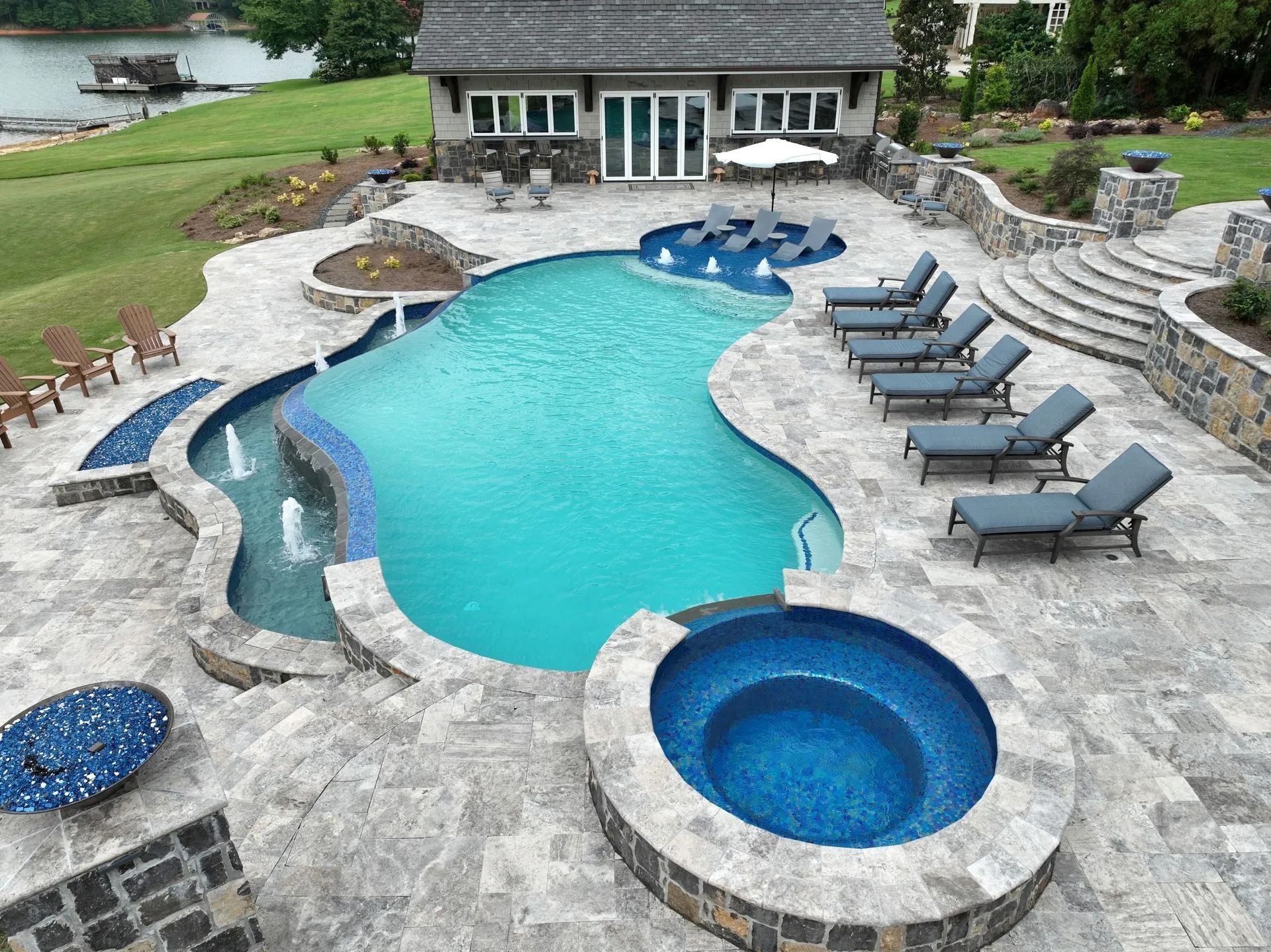 Luxurious backyard pool with spa, lounge chairs, and a small cabana near a lake.