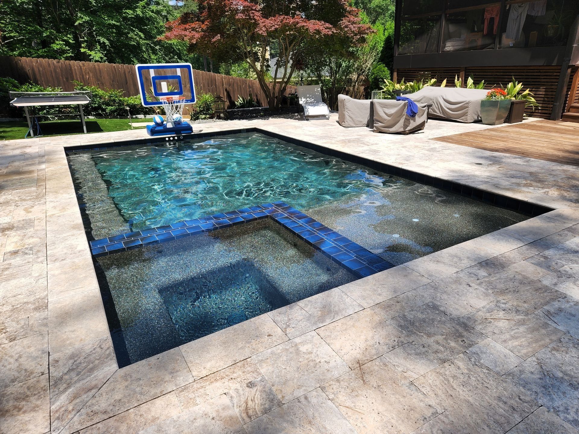 Pool with blue-tiled steps, surrounded by stone patio. Basketball hoop and covered patio furniture in the background.