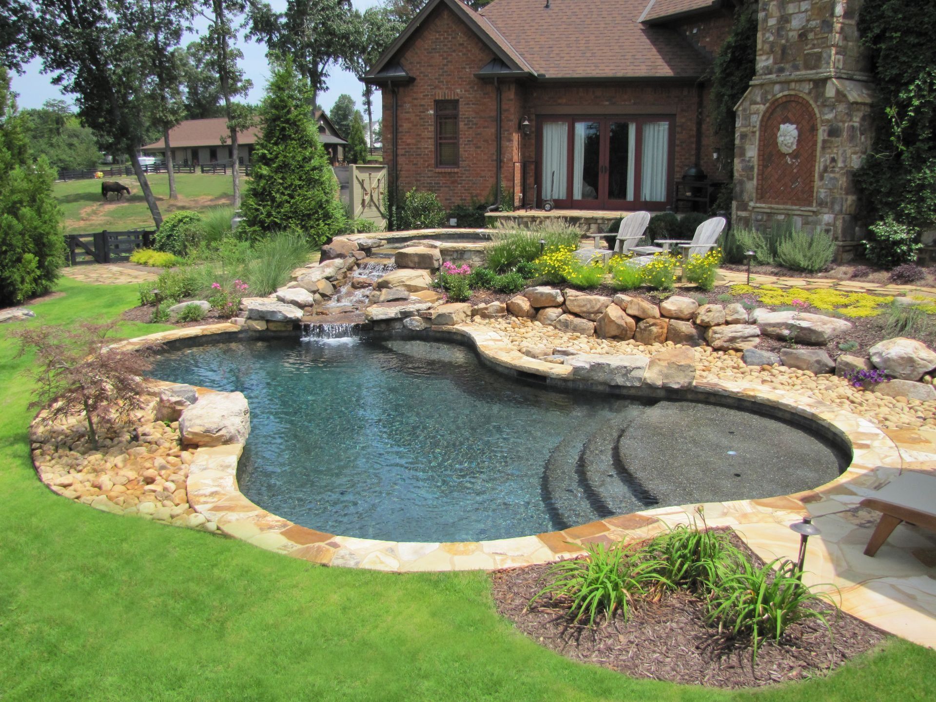 A backyard pool with waterfall, surrounded by landscaping, with a brick house in the background.