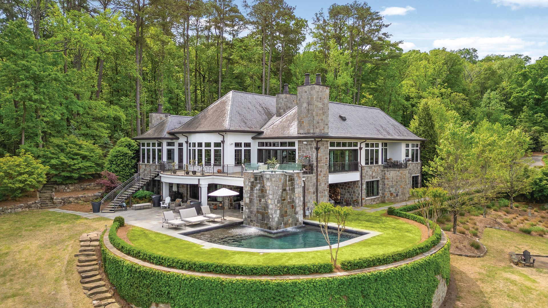 Luxurious stone house with pool and waterfall surrounded by greenery and trees.