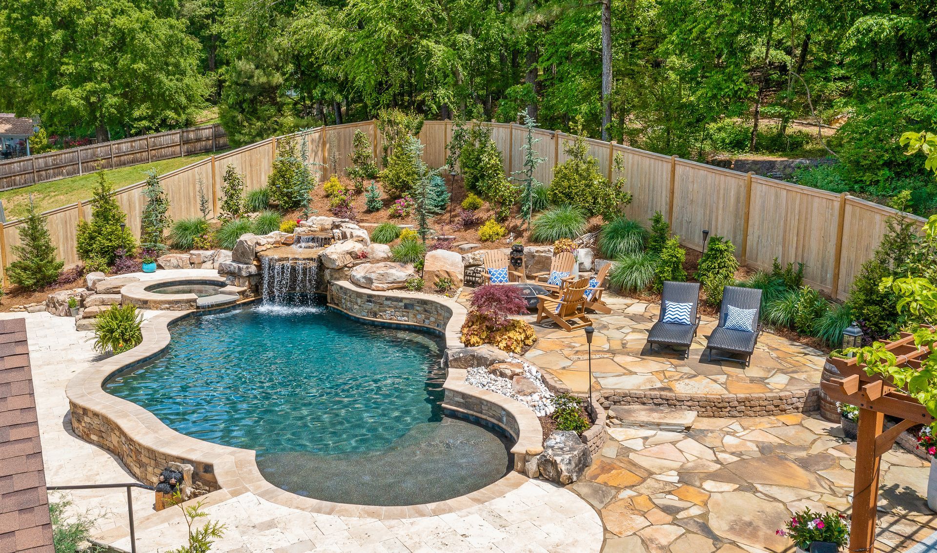 Backyard pool with waterfall, stone patio, lounge chairs, and lush landscaping against a wooden fence.
