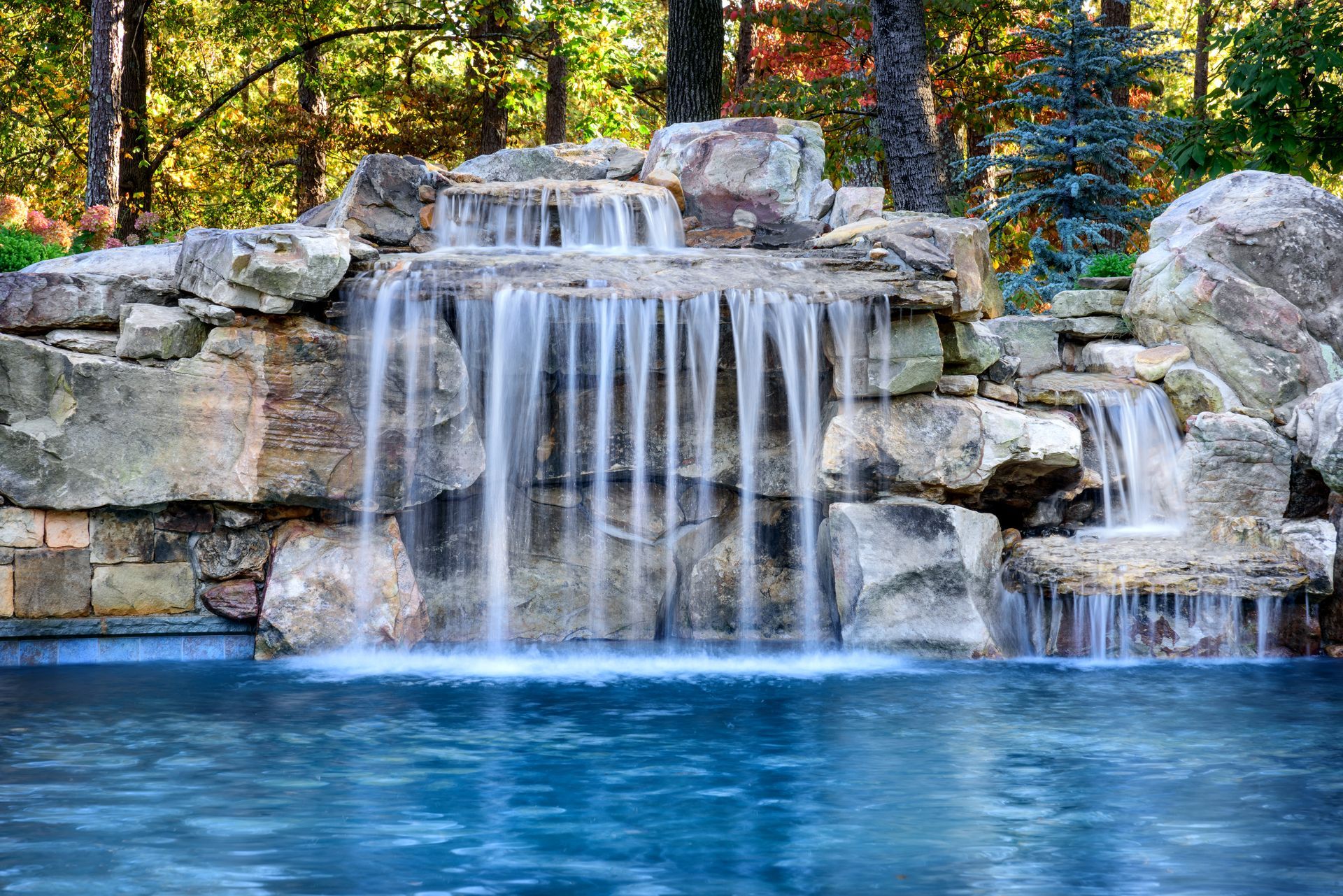 Waterfall cascading into a pool surrounded by rocks and trees. Blue water.