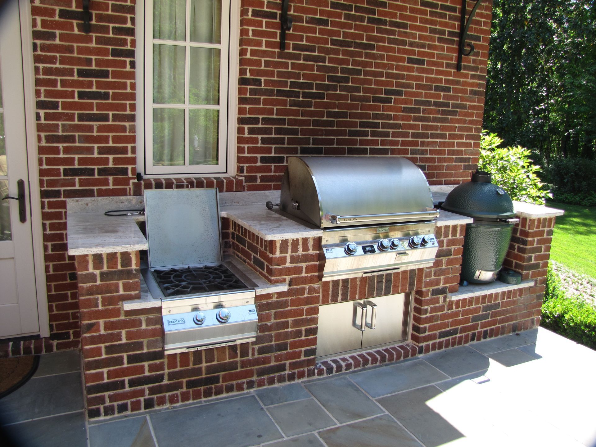 An outdoor kitchen with a brick base and stone countertop, featuring a built-in gas grill, side burner, and ceramic grill.