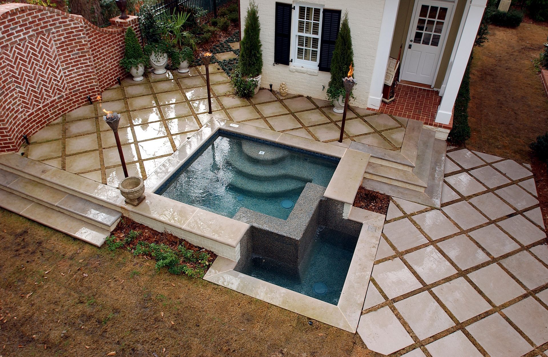 A square swimming pool with a tiered overflow area, set in a light stone patio next to a house with black shutters.