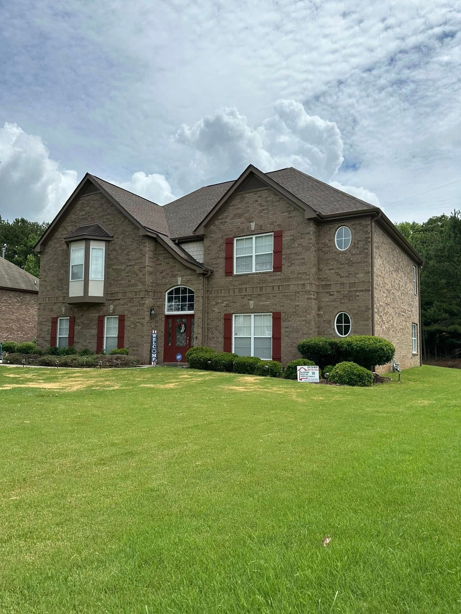 Two-story brick house with dark red shutters, green lawn, cloudy sky background.
