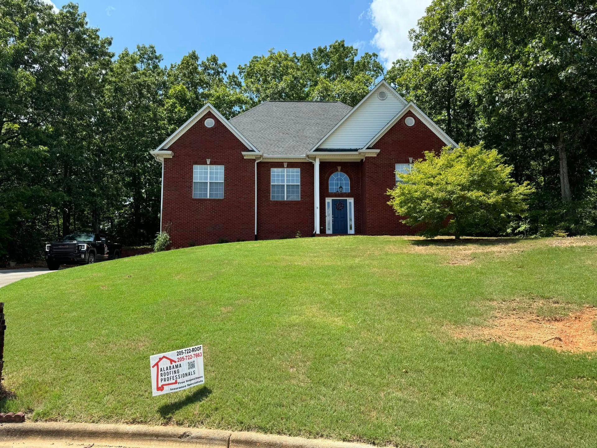 Red brick house on a grassy hill with a sign; surrounded by trees and a blue door.