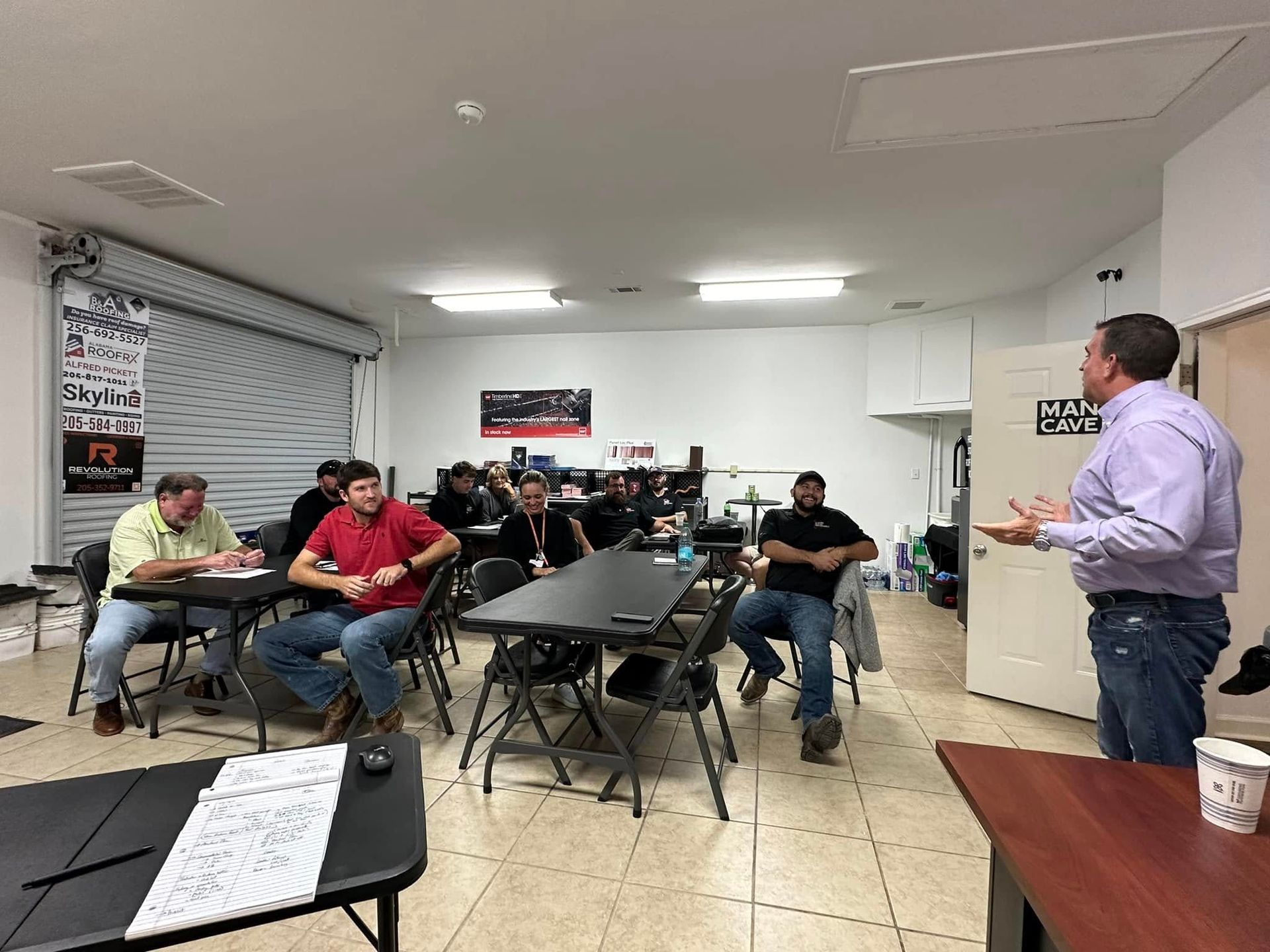 A man lectures to a group seated at tables in a brightly lit room.