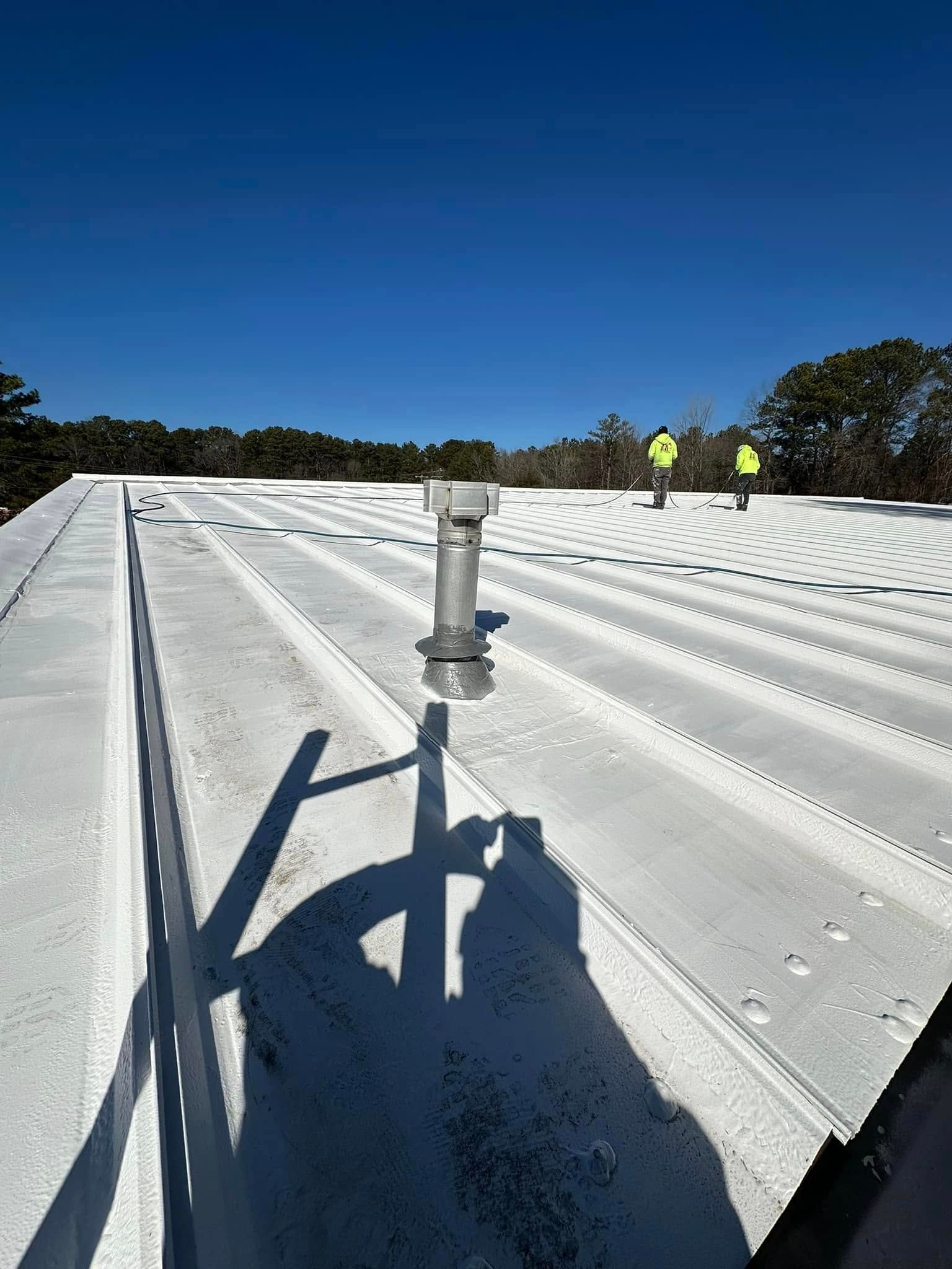 Metal roof with vent pipe; two workers in vests in the background. Bright blue sky.