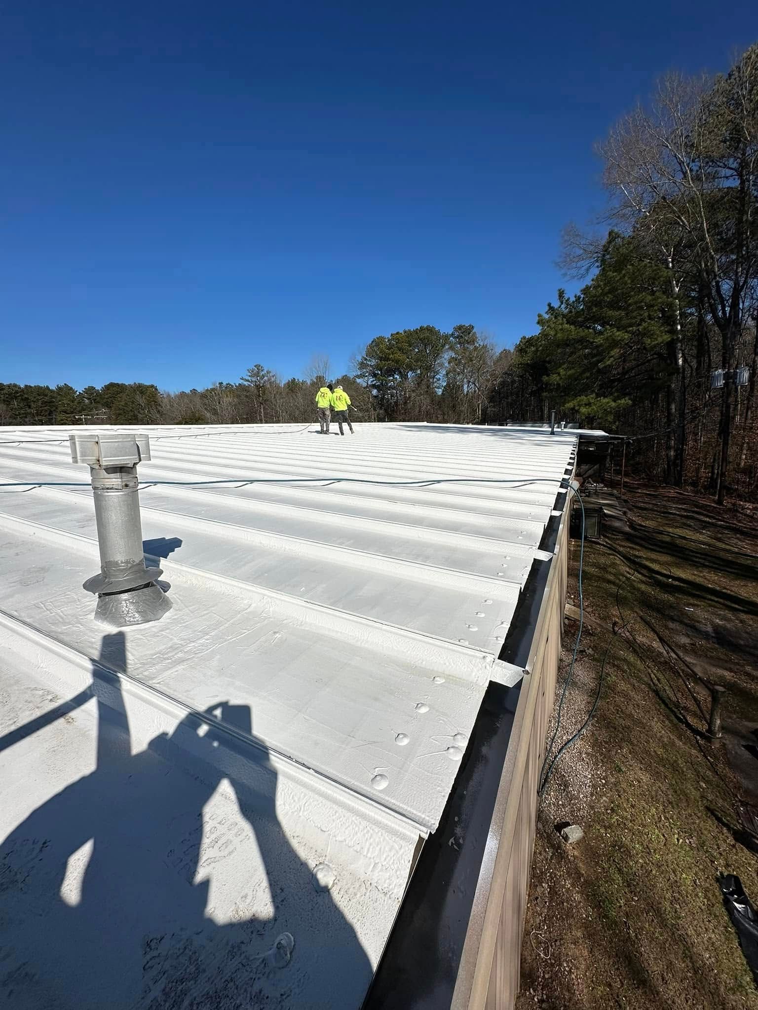 Two workers in yellow vests on a white metal roof on a sunny day. Trees in the background.