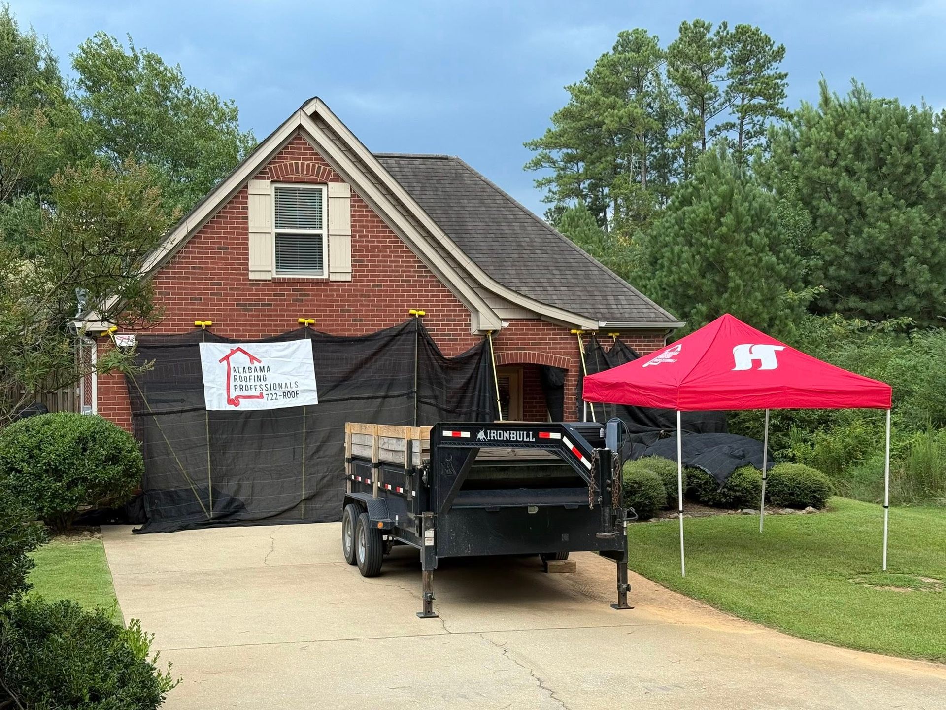 House with construction tarp, trailer, and red tent on the driveway; trees in the background.