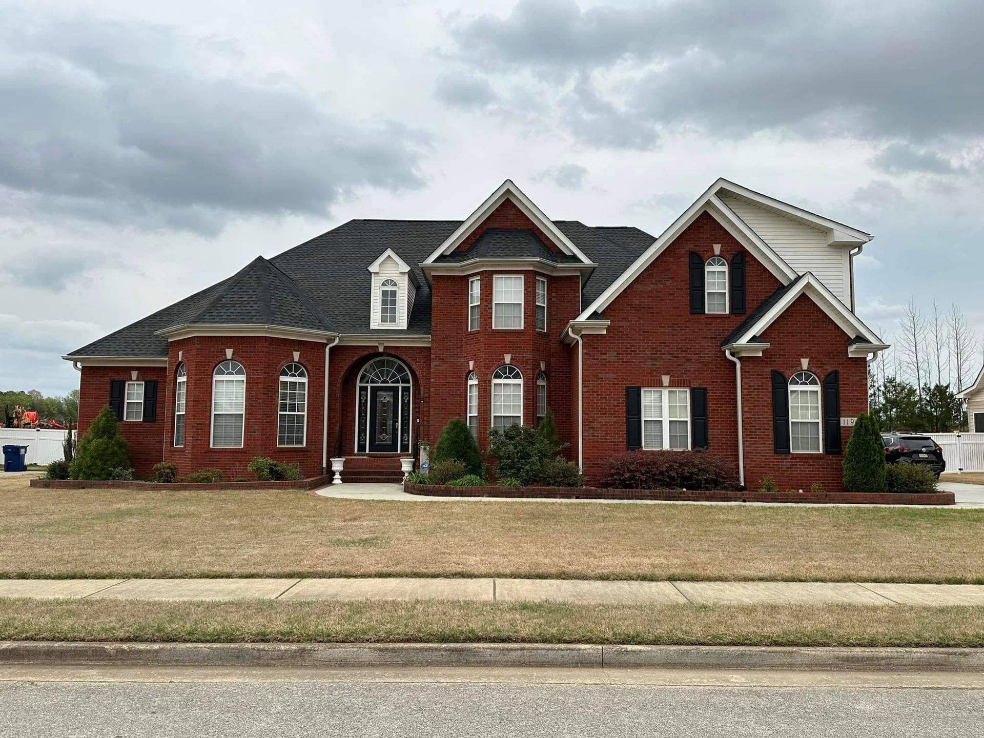 Red brick two-story house with black shutters, dark roof, and a grassy front lawn under a cloudy sky.