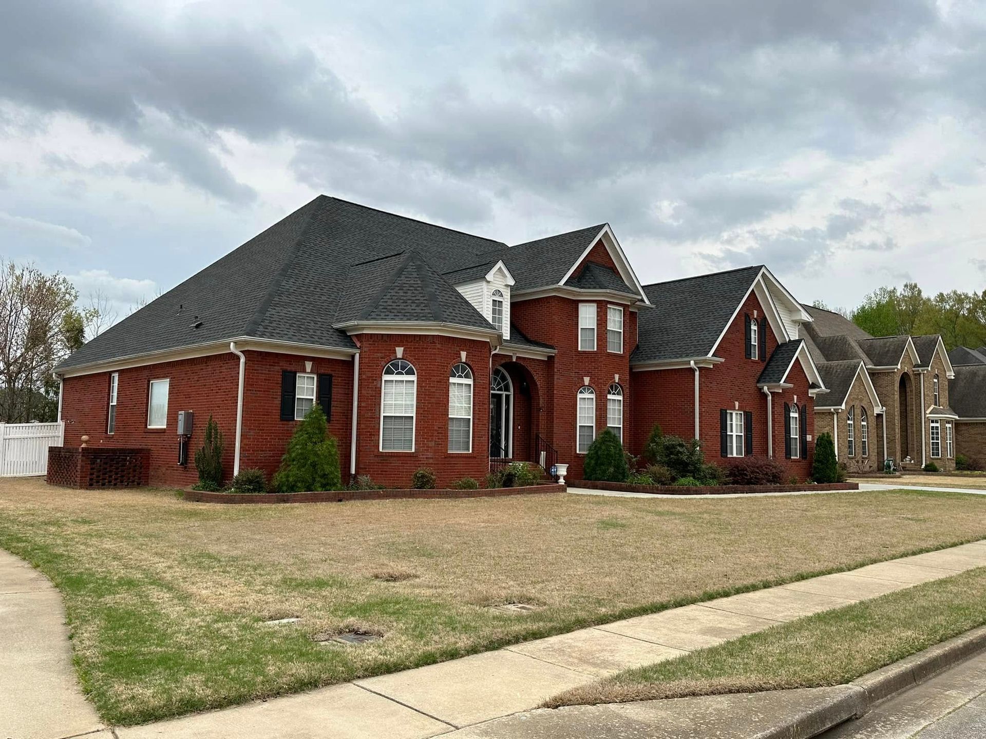 Red brick house with dark roof and brown lawn under a cloudy sky.
