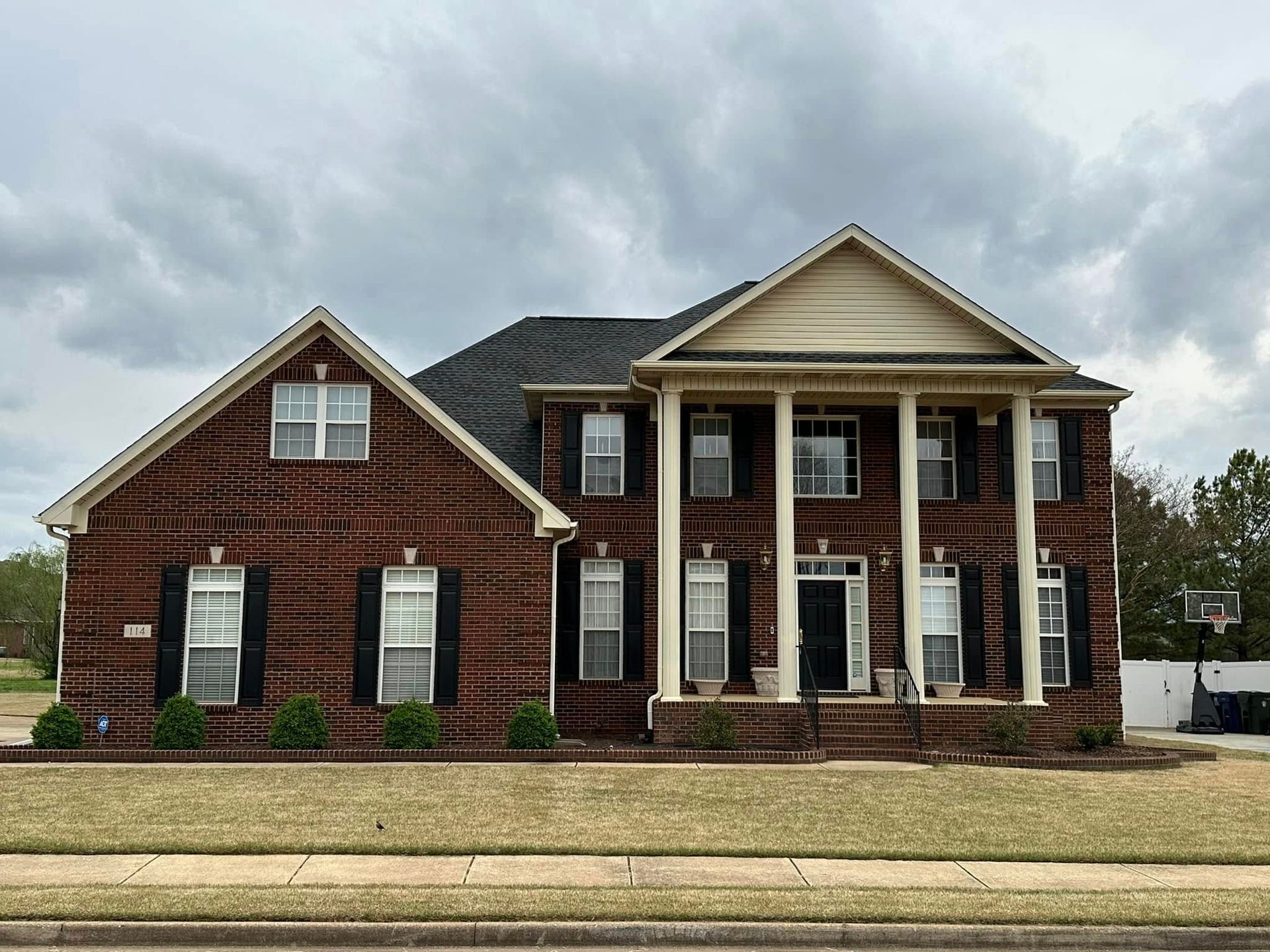 Red brick house with white columns, black shutters, and a dark roof under a cloudy sky.