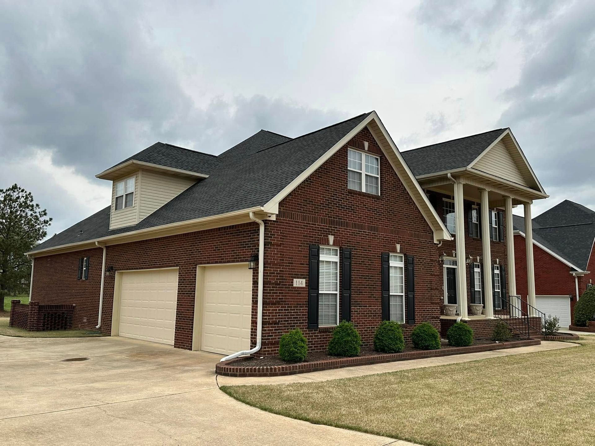 Brick house with columns, two-car garage, and dark roof under a cloudy sky.