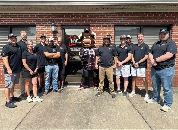 Group of people and a bull mascot posing in front of a brick building with a sign.