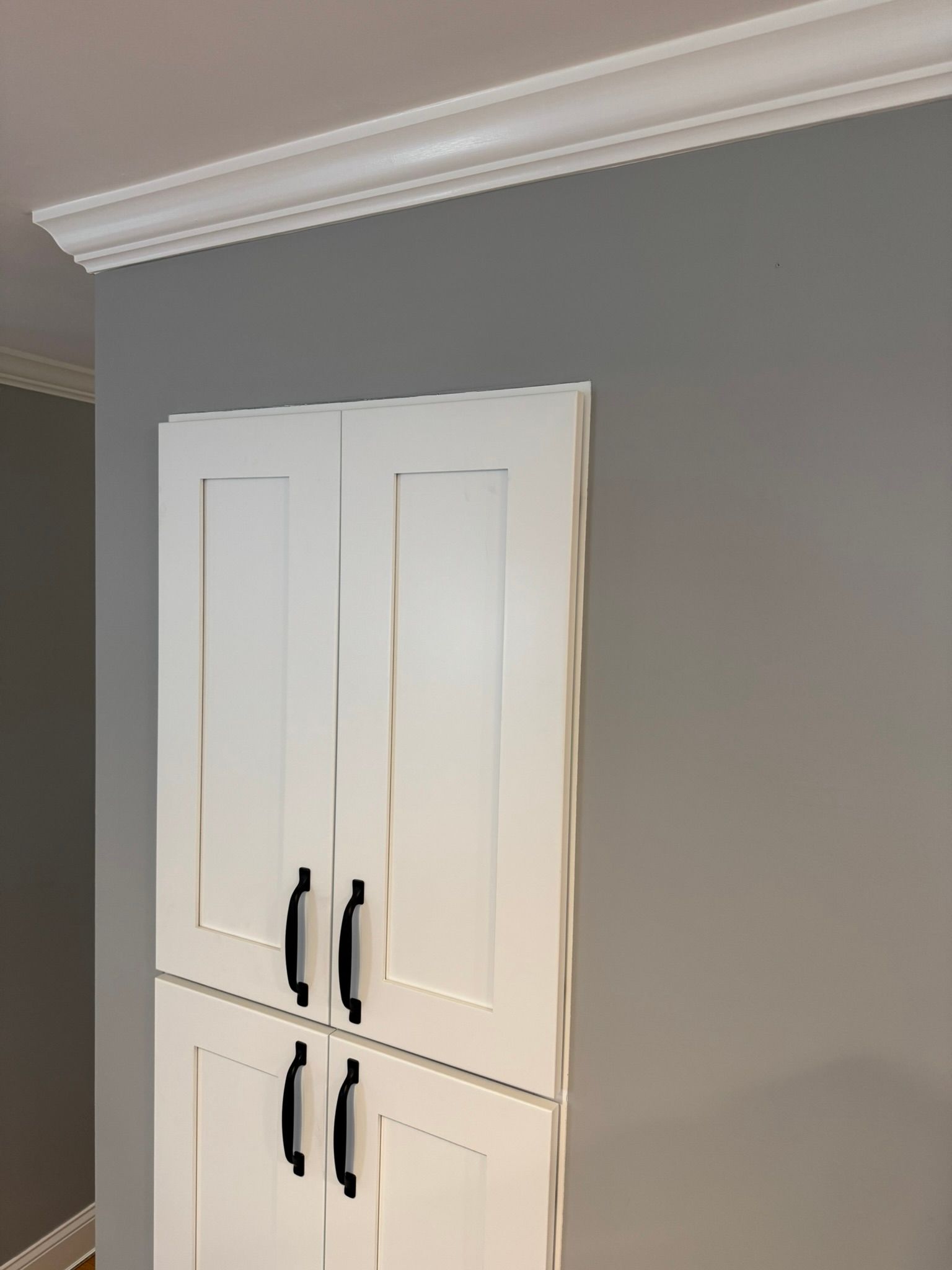 White wall cabinet with black handles mounted on a gray interior wall, beneath crown molding