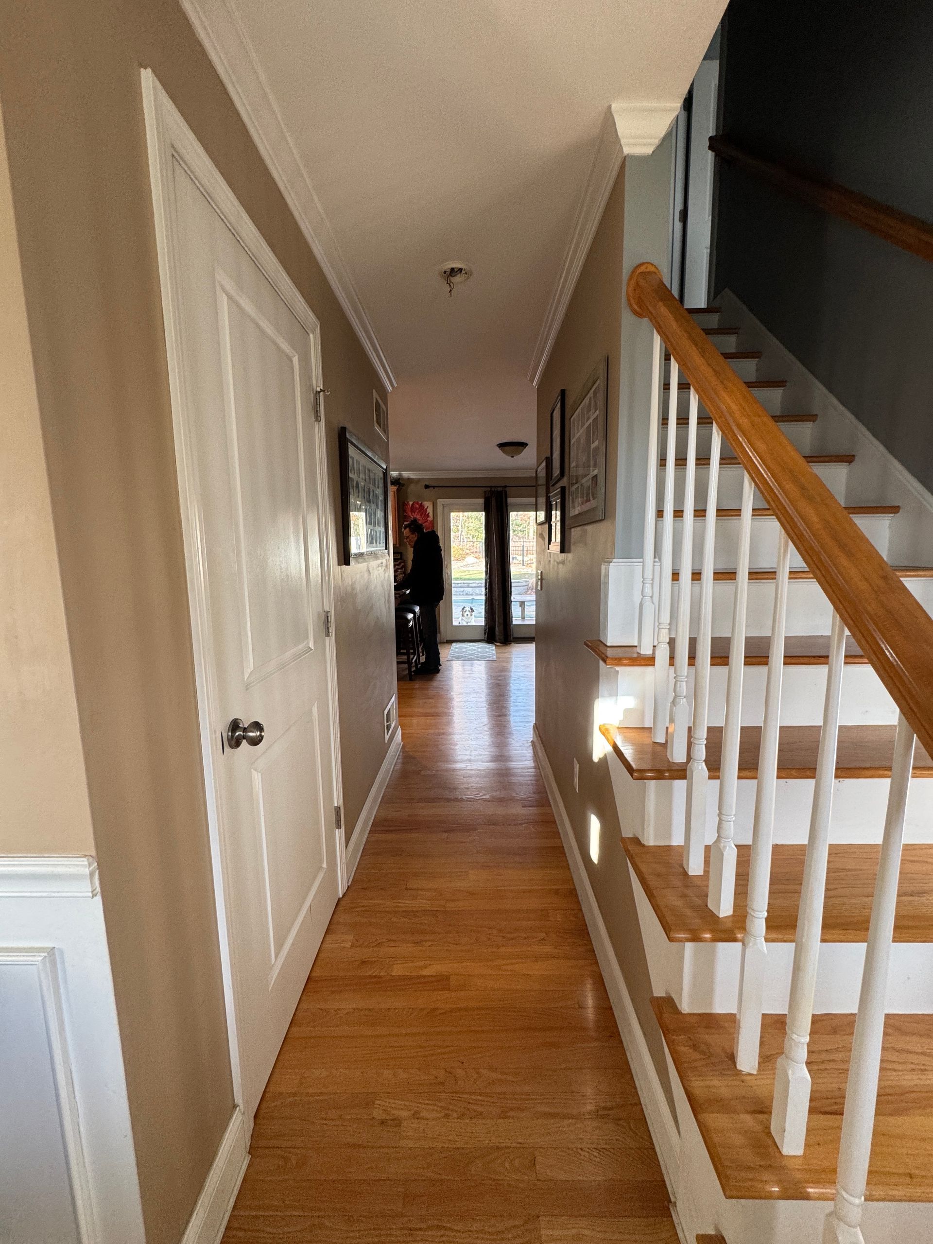 Narrow hallway with hardwood floors, beige walls, and white staircase railing leading to a bright front door.