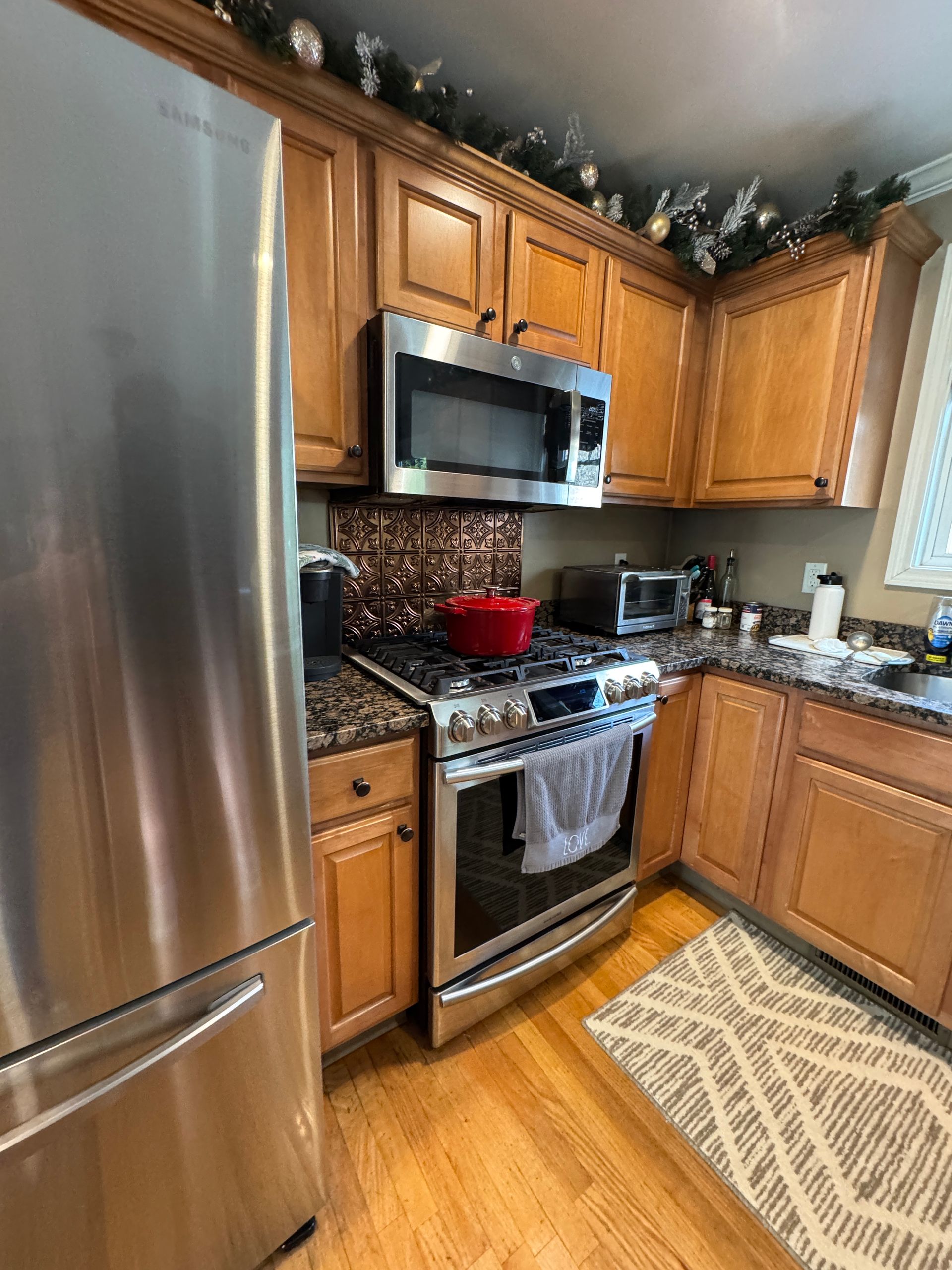 Compact kitchen with stainless steel appliances, wooden cabinets, red pot on stove, and patterned rug