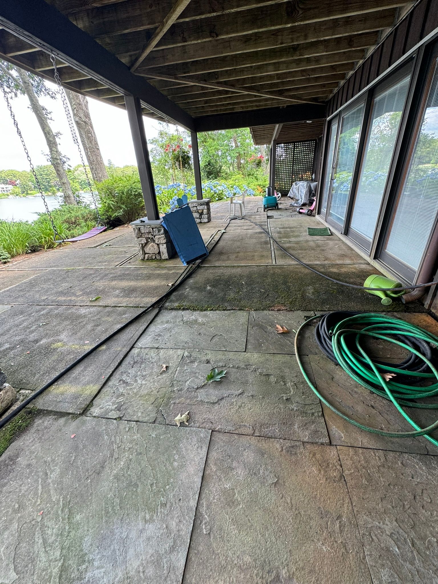 Covered patio with cracked concrete floor, coiled green hose, and garden view beyond the railing