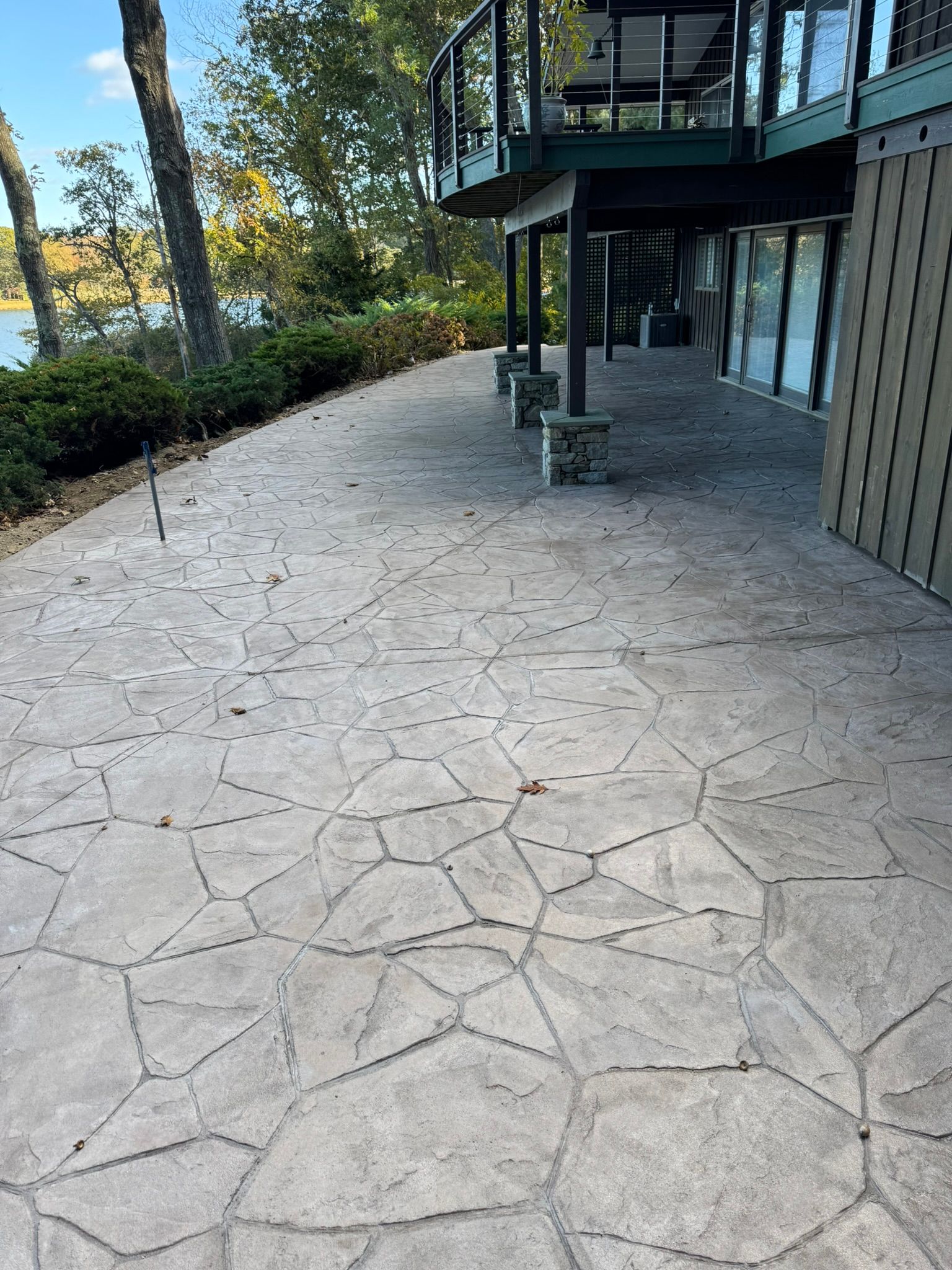 Stone-paved patio beside a modern building with glass doors and a raised balcony, lined with trees and greenery