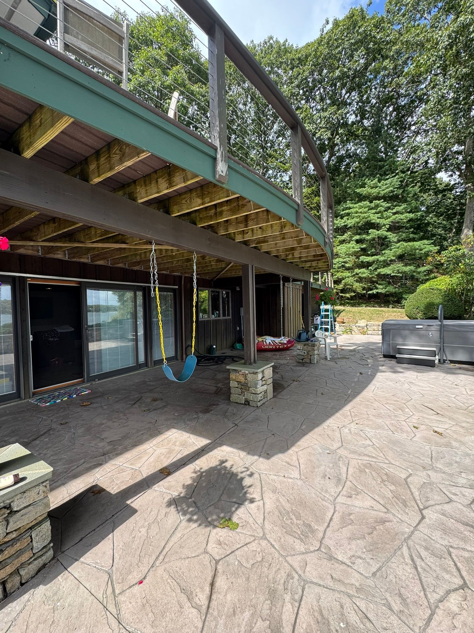 Wooden deck beneath a raised house, with a concrete patio, stairs, and trees in the background.