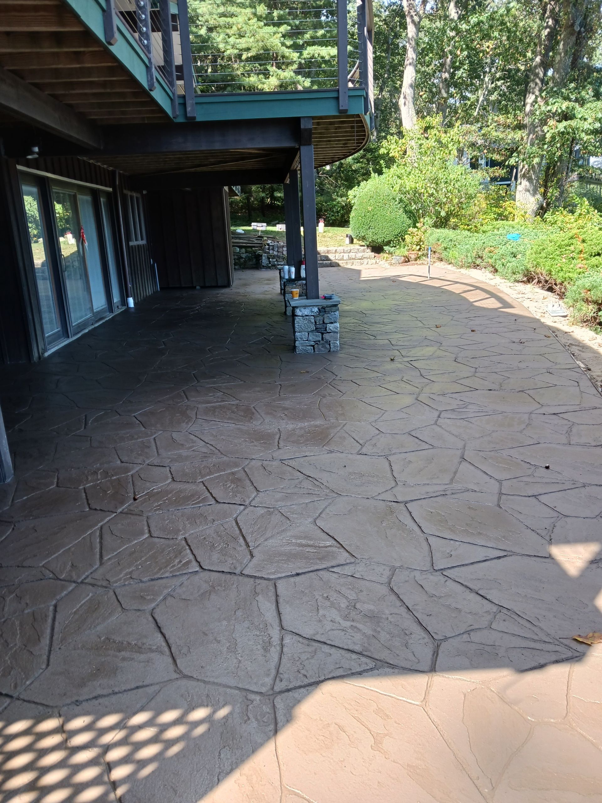 Shaded stone patio beside a wooden deck, with a curved path and sunlit garden edge.