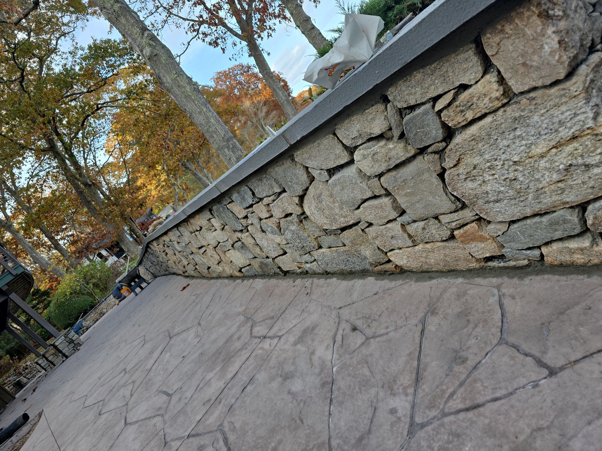Stone wall beside a cracked concrete walkway under autumn trees, viewed at a tilt.