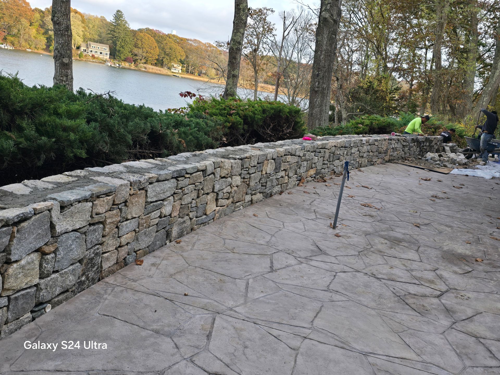 Stone walkway beside a lake, with trees, hedges, and a few people in the distance